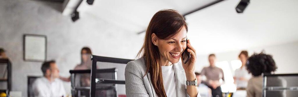 A smiling businesswoman talks on the phone while working on a laptop, representing the personalized service aspect of telecom expense management consulting provided by Chambers Advisory Group.