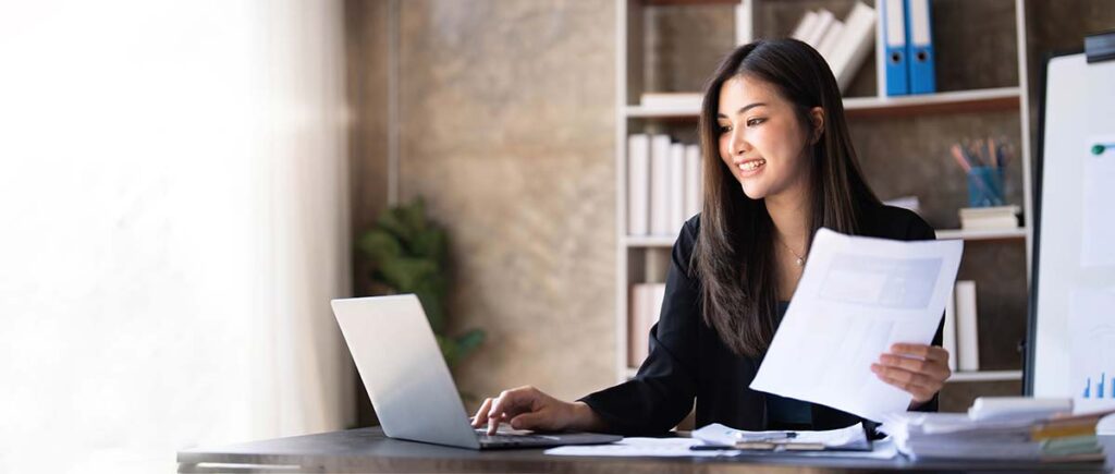 A woman reviewing documents while smiling at her laptop, symbolizing the positive outcomes from Chambers Advisory Group's Telecom Expense Audit services, leading to cost efficiency and streamlined operations.