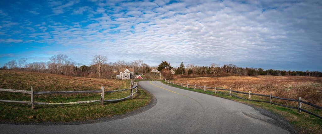 The image shows a winding rural road bordered by wooden fences, with a house in the distance under a partly cloudy sky. The scene conveys a peaceful countryside atmosphere, suggesting a personal touch and community focus.